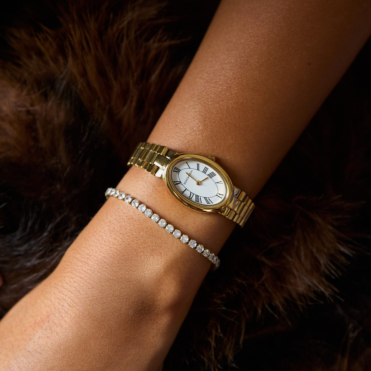 A gold-tone wristwatch with a white dial and Roman numeral markers, worn on a person's wrist alongside a diamond bracelet, set against a dark brown fur background.