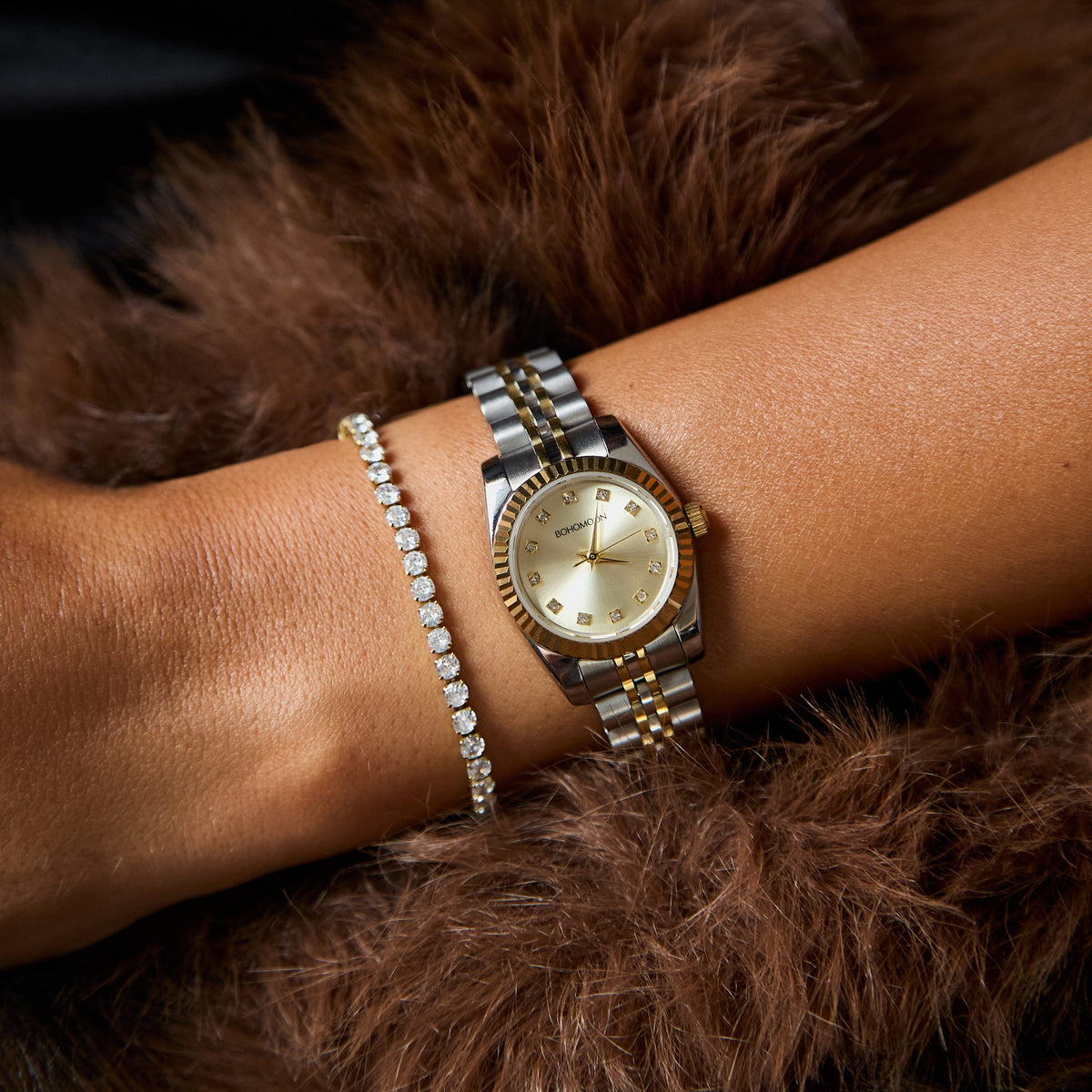 A close-up of a woman's wrist wearing a two-tone gold and silver watch with a gold dial, layered with a diamond-encrusted bracelet, resting against a brown fur surface.